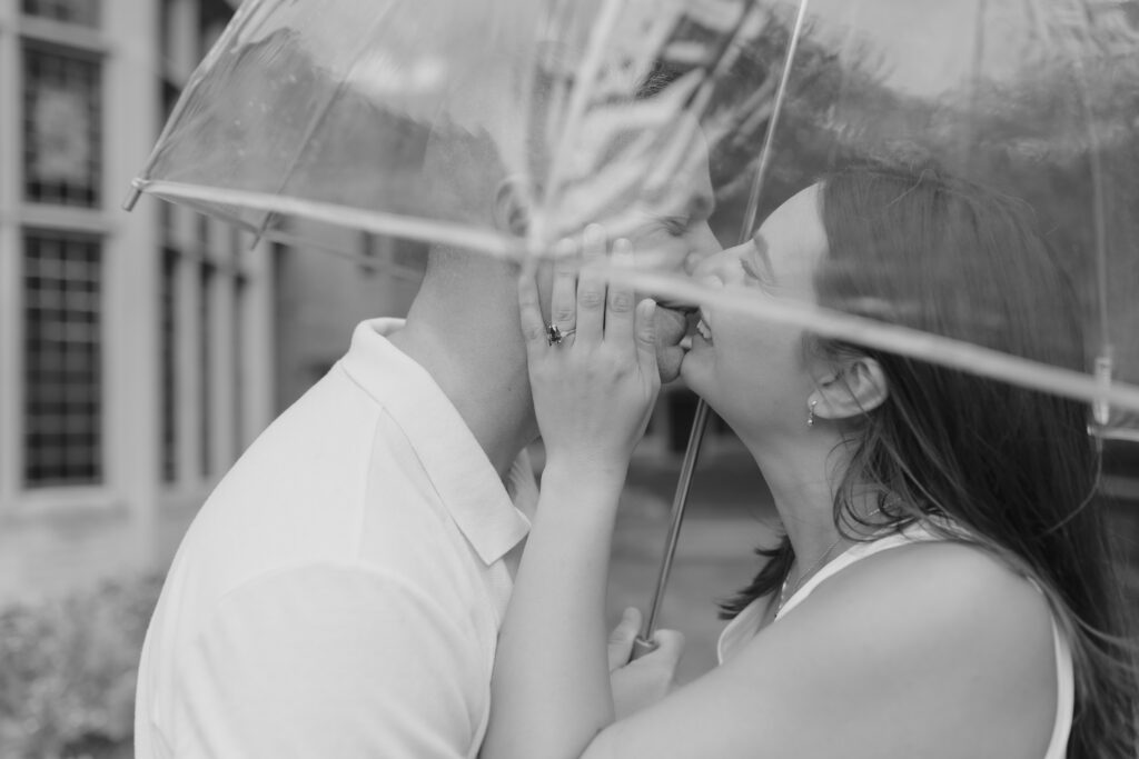 newly engaged couple standing under a clear umbrella kissing